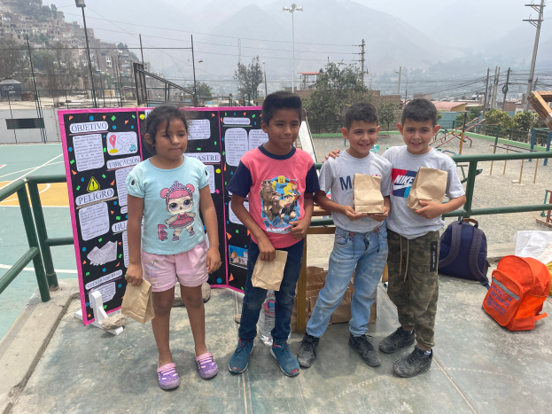 Eine Gruppe von Kindern steht auf einem Basketballfeld, jeder hält eine Papiertüte in der Hand, mit Tüten und Gegenständen rechts daneben und einer Tafel mit Text links daneben, mit einem Zaun, Pfosten, Straßenlaternen, Bäumen, Gebäuden, Hügeln und dem Himmel im Hintergrund.