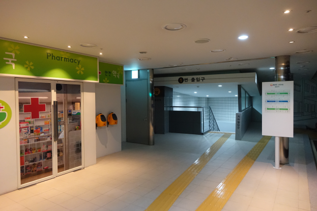 Der Eingang einer Apotheke mit einem "Apotheke"-Schild auf der linken Seite, einer Tafel mit Text auf der rechten Seite und beleuchteten Lichtern auf dem Dach.