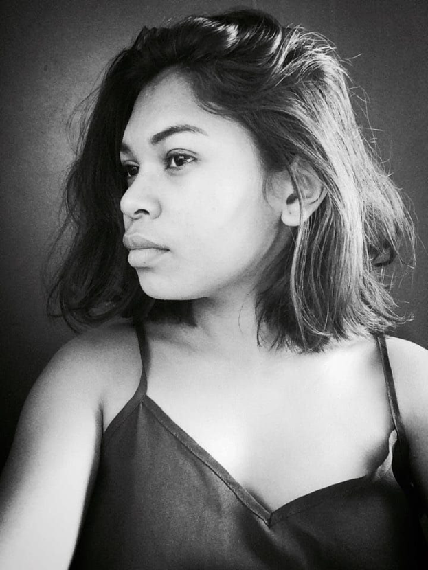 Eine selbstbewusste Frau in einem schwarzen Kleid posiert in einem Schwarz-Weiß-Foto, ihr glatter Bob und das dezente Make-up unterstreichen ihre selbstbewusste Haltung an einer Wand.