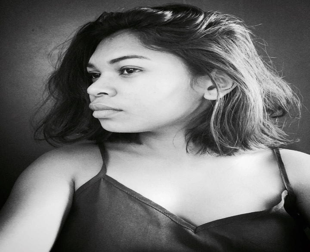 Eine selbstbewusste Frau in einem schwarzen Kleid posiert in einem Schwarz-Weiß-Foto, ihr glatter Bob und das dezente Make-up unterstreichen ihre selbstbewusste Haltung an einer Wand.