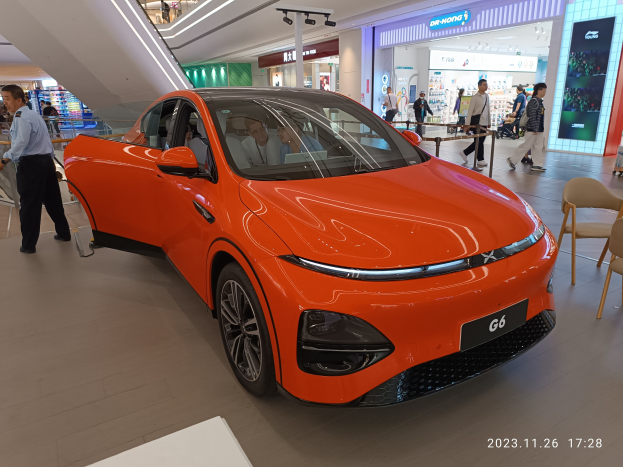 Oranges Elektroauto in einer Ausstellungshalle, umgeben von Menschen, mit Stühlen und einem Wagen rechts und Geschäften im Hintergrund.