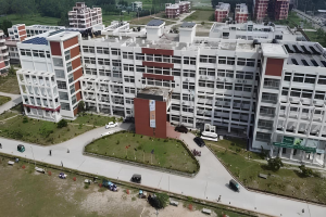 Luftaufnahme eines großen Krankenhausgebäudes mit Solarpaneelen auf dem Dach, umgeben von Bäumen, Gras, Fahrzeugen, Menschen, Strommasten und einer Tafel, die ein Solarprojekt anzeigt.