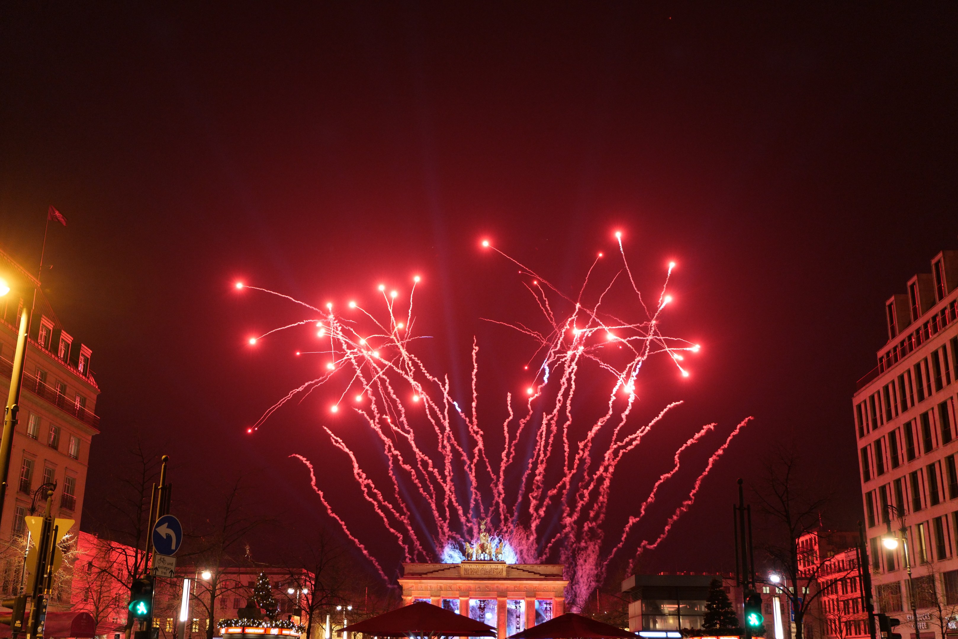 Eine belebte Stadtstraße an einem Silvesterabend in Berlin, mit Gebäuden, Bäumen, Laternen, Ampeln, Schildern, Zelten, Menschen und einem prächtigen Feuerwerk am Himmel.