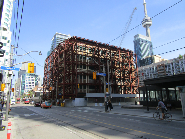 Eine belebte Stadtstraße mit einem großen Baugerüst im Hintergrund, mit Fahrzeugen, einem Radfahrer, Fußgängern, Ampeln, Pfosten mit Schildern, einem Kran und dem Himmel.