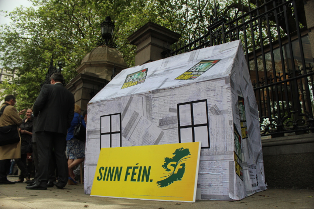 Ein Zelt und ein Werbeplakat sind auf dem Boden platziert, während eine Menge daneben steht. Im Hintergrund sind ein Eisenzaun, Säulen, Straßenlaternen, Bäume und der Himmel zu sehen.
