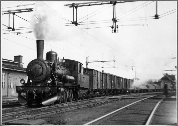 Schwarzes und weißes Foto eines Dampflokomotivs auf Bahnschienen mit Rauch aus dem Motor, ein Gebäude auf der linken Seite und Strommasten mit Drähten im Hintergrund.