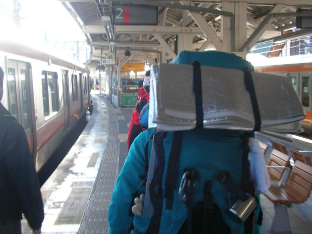 Ein Bahnhof mit Menschen neben einem Zug, ein Stand in der Mitte, ein weiterer Zug auf der rechten Seite und eine Bank in der rechten unteren Ecke.