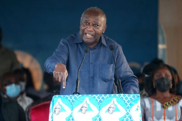 Ein Mann in einem blauen Hemd spricht an einem Pult mit zwei Mikrofonen und schaut auf eine sitzende Menge, einige tragen Masken, mit einer Wand im Hintergrund.