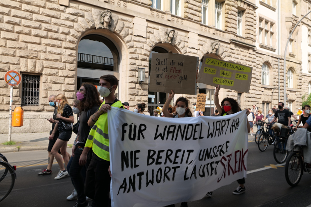 Eine Gruppe von Menschen marschiert auf einer Straße, einige halten Protestschilder und andere fahren Fahrräder, während im Hintergrund ein historisches Gebäude mit Fenstern, Bögen, Säulen und Skulpturen zu sehen ist.