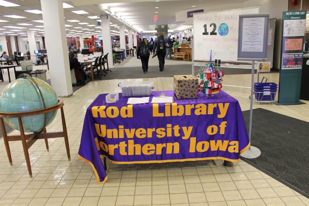Eine Ausstellung mit einem Tisch, auf dem verschiedene Landesflaggen, eine Box, Papiere und ein Banner sowie links daneben eine Weltkugel, rechts ein Anschlagbrett mit einigen Menschen in der Nähe und mehrere Tische und Stühle im Hintergrund zu sehen sind.