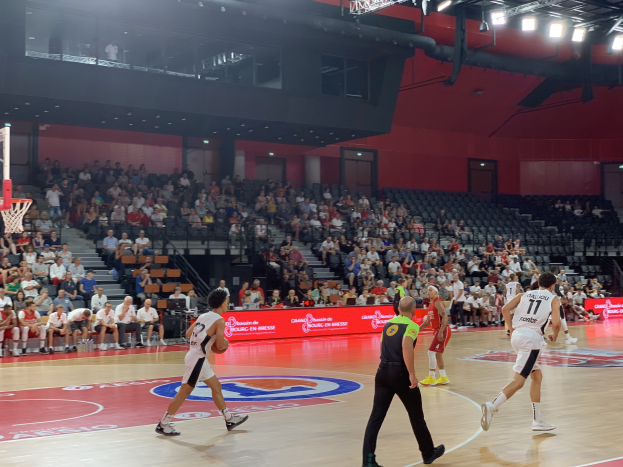 Eine Gruppe von Menschen, die Basketball in einer Halle mit einem Netz auf der linken Seite spielen, Zuschauern im Hintergrund und Deckenbeleuchtung.