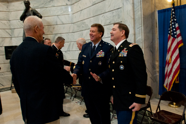 Menschen schütteln sich die Hände in einem Raum mit Stühlen, einer Statue neben einer Wand, einer Flagge und einem Vorhang auf der rechten Seite.