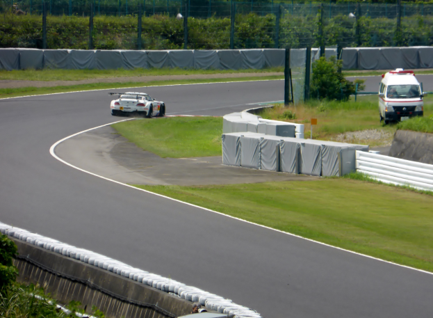 Ein Rennwagen fährt eine Strecke mit einem Polizeiwagen im Hintergrund, umgeben von Gras, Pflanzen, Bäumen und Zaun, mit einem Fahrzeug auf der rechten Seite und Text unten.