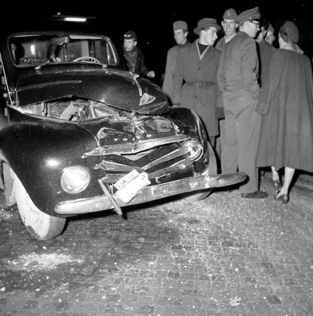 Eine Schwarz-Weiß-Szene eines beschädigten Autos am Straßenrand, mit einer Gruppe besorgter Menschen drumherum.