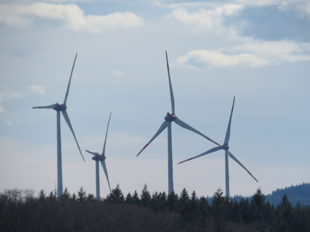 Eine Gruppe von Windkraftanlagen auf einem Feld umgeben von Bäumen, mit Hügeln im Hintergrund und Wolken am Himmel.