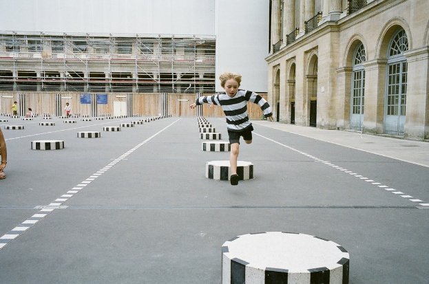 Ein Spielplatz mit vier Kindern, eines rennt auf dem Boden mit schwarzen und weißen Betonstufen, umgeben von einem Schulgeb├Ąude.