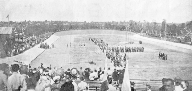 Schwarz-weißes Foto von Zuschauern bei einem Pferderennen in einem Stadion, einige sitzen auf Bänken und andere stehen oder reiten Pferde, mit Bäumen und einem klaren Himmel im Hintergrund.