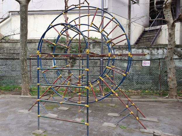 Spielplatz mit großem Metall-Klettergerüst, umgeben von Bäumen und einer eingefassten Wand, mit Treppen mit Geländern, die zu einem Gebäude mit Fenstern und Rohren führen, einem Schild an einem Pfahl und grasbewachsenem Boden.