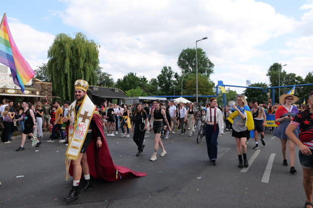 Eine Gruppe von Menschen bei der 2018er Christopher Street Day Parade mit einer Regenbogenflagge und Musikinstrumenten, umgeben von Laternenmasten, Bäumen, Häusern und einem bewölkten Himmel.