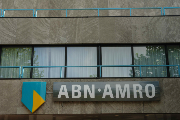Ein Gebäude mit einer Tafel, auf der "ABN AMRO" steht, mit einer Tafel, die anzeigt, dass es der Hauptsitz ist, Geländern und Glasfenstern mit Vorhängen, sowie einer Notiz über eine Strafe von 1,5 Milliarden Dollar durch die Europäische Union.