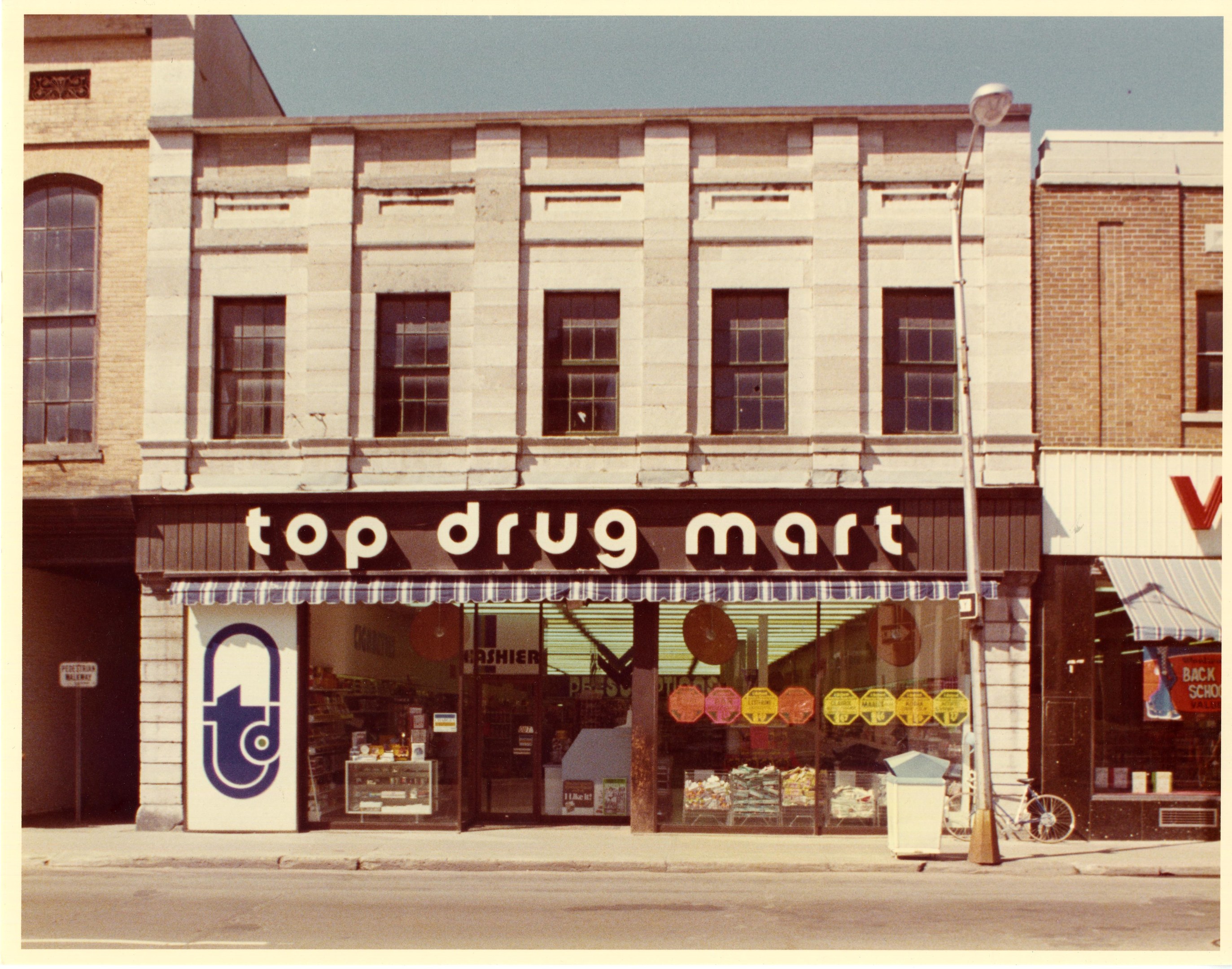 Eckladen "Top Drug Mart" mit Fenstern, einem Schild, einem Straßenschild, einem geparkten Fahrrad, einem Müllcontainer und einem bewölkten Himmel.