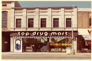 Eckladen "Top Drug Mart" mit Fenstern, einem Schild, einem Straßenschild, einem geparkten Fahrrad, einem Müllcontainer und einem bewölkten Himmel.