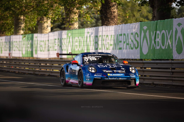 Blauer Porsche 911 GTS auf einer Rennstrecke mit einem Zaun und Bannern im Hintergrund, Bäume und einen klaren blauen Himmel.