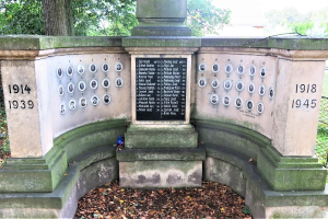 Ein Holocaust-Denkmal in einem jüdischen Friedhof in Berlin, mit einer Text- und Nummernwand an seiner Mauer, umgeben von Bäumen, einem Zaun und trockenen Blättern auf dem Boden.