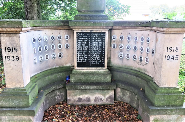 Ein Holocaust-Denkmal in einem jüdischen Friedhof in Berlin, mit einer Text- und Nummernwand an seiner Mauer, umgeben von Bäumen, einem Zaun und trockenen Blättern auf dem Boden.