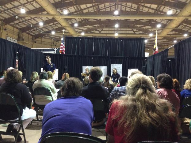 Eine Gruppe von Menschen, die auf Stühlen sitzen, mit einer Person, die an einem Rednerpult steht, in einem Raum mit Bannern, blauen Vorhängen, Flaggen und Deckenleuchten.