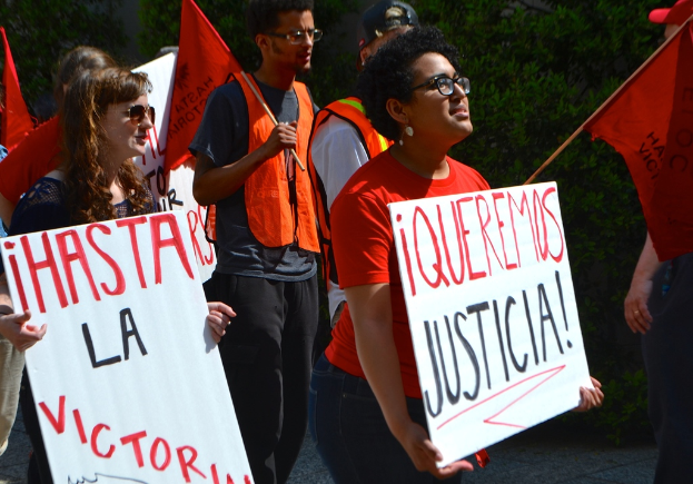 Mehrere Personen in orangefarbenen Kleidern stehen mit wei√üen Schildern und orangefarbenen Fahnen, mit einer Frau in einem schwarzen Kleid im Vordergrund und gr√ľnen B√§umen im Hintergrund.