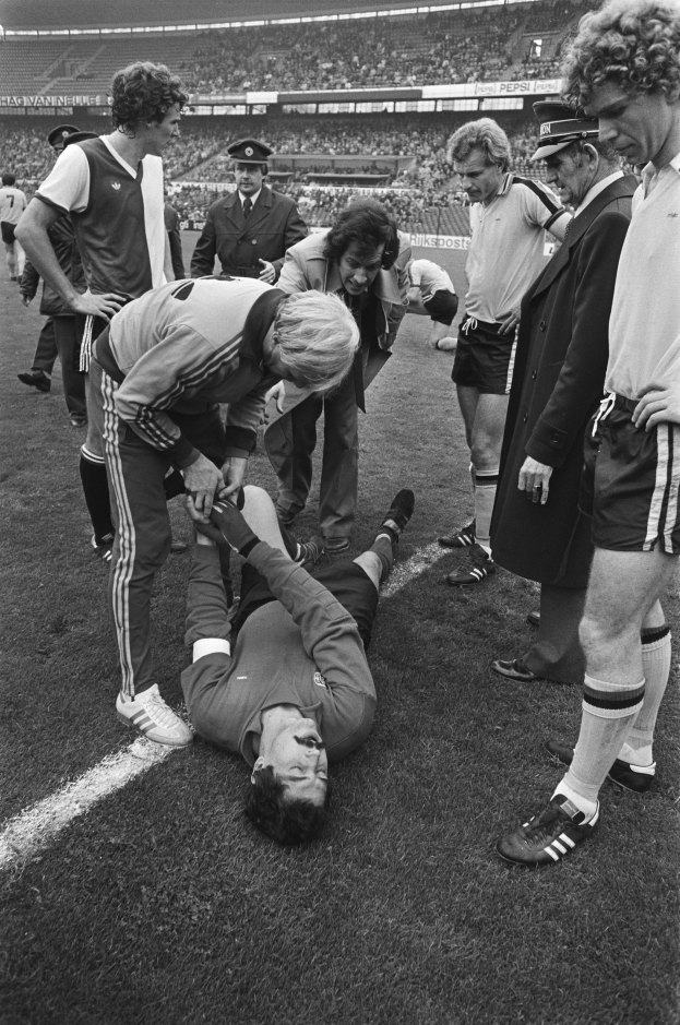 Schwarzes und weißes Bild eines Fußballspielers, der auf dem Boden liegt und von einer Gruppe von Männern umgeben ist, während eine Stadionmenge zusieht.