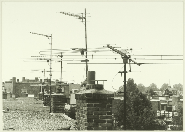 Eine Schwarz-Weiß-Stadtlandschaft mit zahlreichen Gebäuden, Bäumen und Fernsehantennen auf einem Dach.