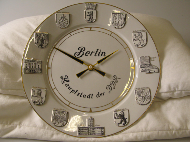 Eine Uhr mit dem Wort 'Berlin' darauf steht auf einem weißen Kissen an einer Wand im Hintergrund.