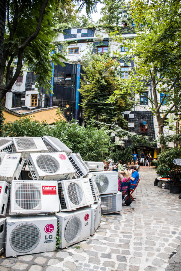 Luftkühler am Boden links unten, eine Spur rechts daneben, Menschen auf Stühlen neben der Spur, mit Gebäuden, Bäumen und Pflanzen darüber.