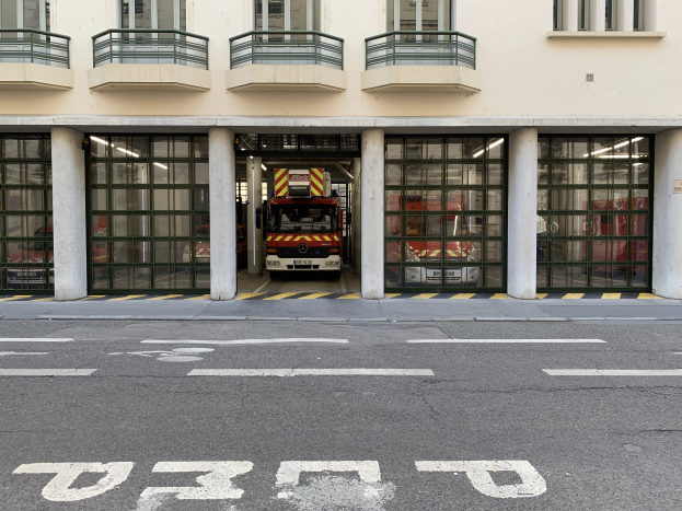 Feuerwache mit Feuerwehrauto vor dem Gebäude, mit Fenstern, Geländern, Säulen und Glastüren, sowie einer Straße unten.