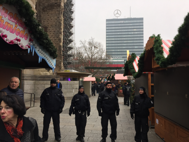 Eine Gruppe von Polizisten in schwarzen Uniformen steht vor einem festlichen Weihnachtsmarkt in Berlin, Deutschland, mit Gebäuden, Bäumen und einem klaren blauen Himmel im Hintergrund und einer Tafel mit Text auf der linken Seite des Bildes.
