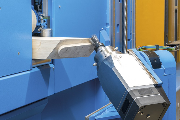 Eine CNC-Fräsmaschine schneidet ein Holzstück in einer Fabrikumgebung, umgeben von Kabeln und Gegenständen, mit einer Wand im Hintergrund.