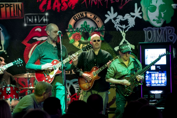 Drei M├Ąnner, die auf einer B├╝hne Gitarren spielen, vor einem Publikum und einer Wand mit Graffiti im Hintergrund.