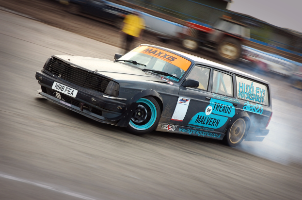 Ein Auto fährt eine Rennstrecke entlang und produziert dabei Rauch, umgeben von anderen Fahrzeugen und Menschen.