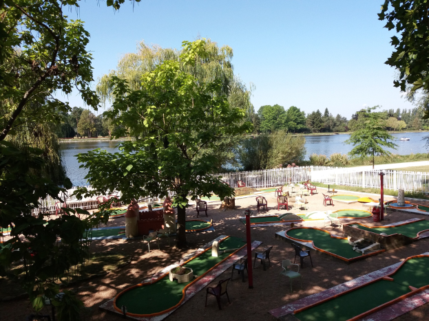 Minigolfplatz in einem Park mit grünem Gras, Bäumen, einem Zaun, verstreuten Stühlen und Tischen, einem Gewässer mit einem Boot und sichtbarem Himmel.