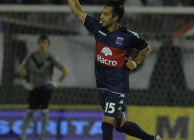 Ein Fußballspieler feiert nach einem Tor auf dem Boden liegend, mit jubelnden Zuschauern und einer Person im Hintergrund, alles vor einem leicht unscharfen Hintergrund.