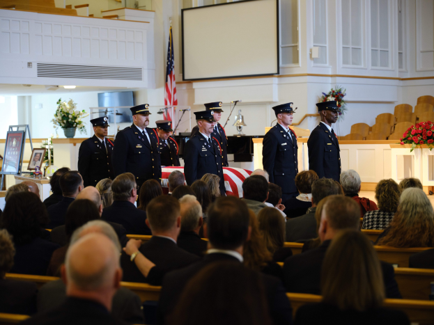 Eine Gruppe uniformierter Personen mit Mützen, die vor sitzenden Trauernden bei einer Beerdigung stehen, mit Blumenbouquets, einer Fahne, einem Bildschirm und einer Wand im Hintergrund.