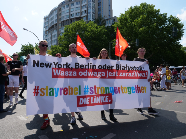 Eine Gruppe von Menschen, die eine Straße in Berlin, Deutschland, entlanggehen und ein Transparent mit der Aufschrift "Stay Rebel Stronger Together" halten, mit Bäumen, Laternenpfählen und Gebäuden an der Straße und einem bewölkten Himmel.