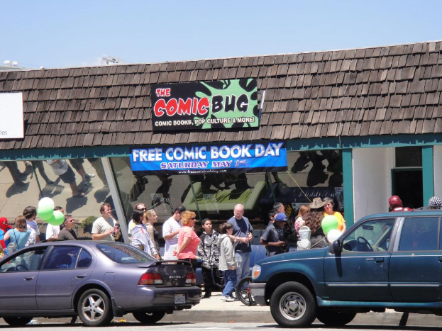 Gruppen von Menschen stehen vor einem Gebäude mit Glastüren und einem Dach, zwei Autos auf der Straße, grüne Ballons, eine Fahne und ein Namensschild auf dem Dach, unter einem klaren Himmel.