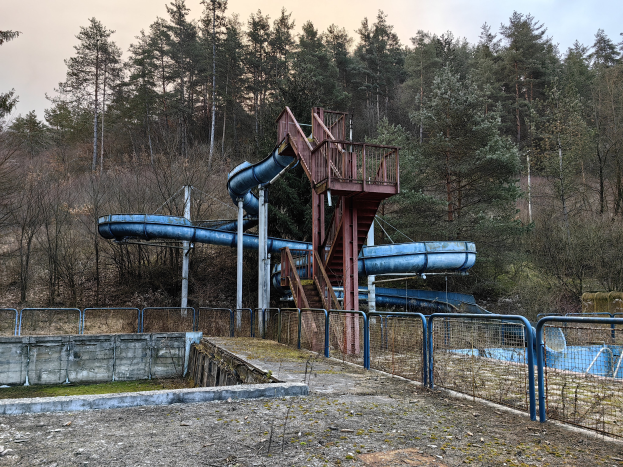 Ein verlassenes Wasserpark mit einer blauen Wasserrutsche in der Mitte, umgeben von Geländern, Treppen und einer Wand, mit Bäumen und einem klaren blauen Himmel im Hintergrund und Text am unteren Bildrand.