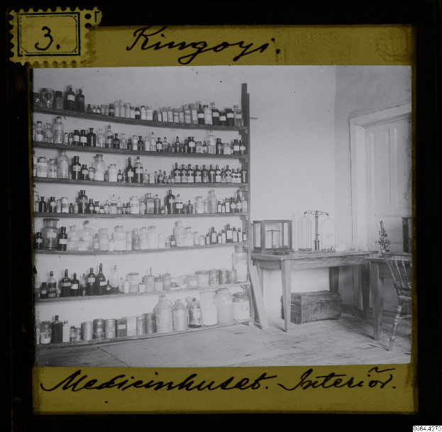 Schwarz-weißes Foto einer alten Apotheke mit Regalen voller Flaschen, einem Tisch mit Gegenständen, einem Stuhl, einer Tür und einer Wand, mit Text unten.