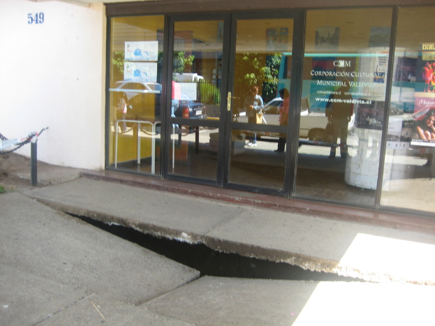 Eine zerbrochene Fläche im Vordergrund mit einer Glastür, Wand und einem Pfahl im Hintergrund; die Spiegelung in der Glasscheibe zeigt fahrende Fahrzeuge und einige stehende Personen.
