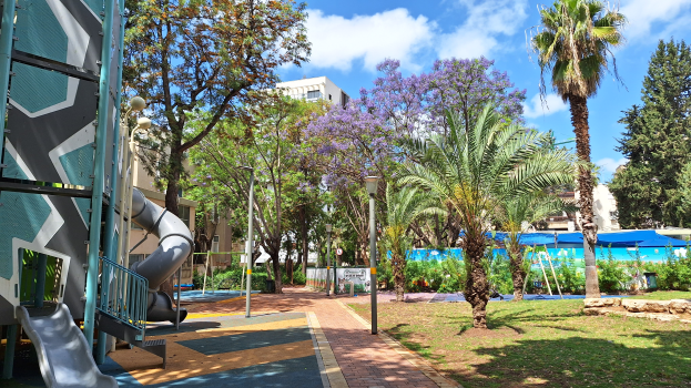 Ein Spielplatz mit einer Rutsche und Palmen, umgeben von Gebäuden, Straßenlaternen, Gittern, Pflanzen, Bäumen, Gras, Felsen und einem bewölkten Himmel im Hintergrund.