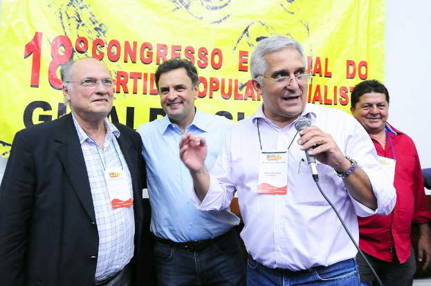 Ein Mann spricht in ein Mikrofon vor einem gelben Vorhang mit der Aufschrift "18 Grad Kongress o", w├Ąhrend andere M├Ąnner im Hintergrund stehen.